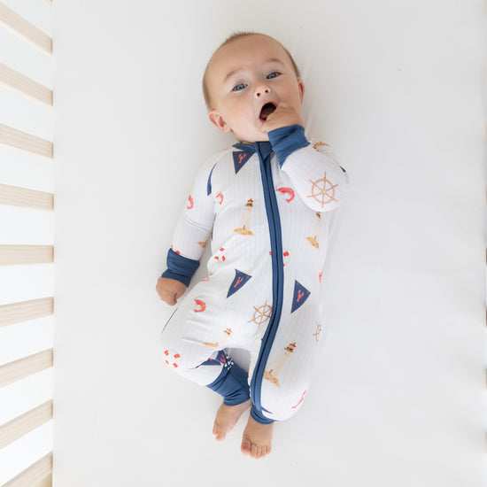A baby lying on their back on a white mattress, wearing a white zip-up sleeper with a blue zipper, blue cuffs and collar, and a nautical print of ships wheels, anchors, and sailboats.