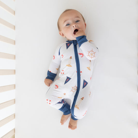 A baby lying on their back on a white mattress, wearing a white zip-up sleeper with a blue zipper, blue cuffs and collar, and a nautical print of ships wheels, anchors, and sailboats.
