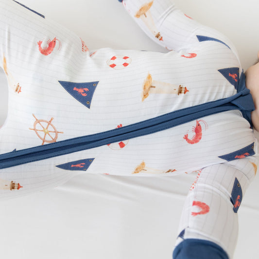 Infant model wearing a white zip-up sleeper with a blue zipper, blue cuffs and collar, and a nautical print of ships wheels, anchors, and sailboats.
