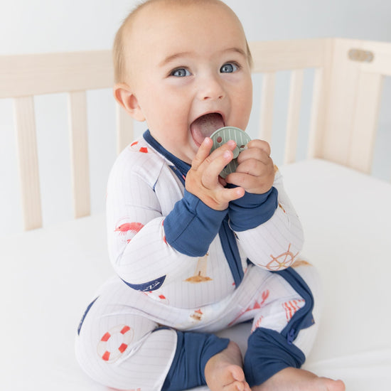Infant sitting in a crib wearing a white zip-up sleeper with a blue zipper, blue cuffs and collar, and a nautical print of ships wheels, anchors, and sailboats.