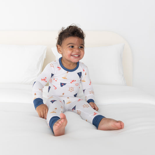 A smiling toddler sits on a white bed wearing a long-sleeve pajama set with a nautical print. The pajamas are white with navy blue cuffs and collar, decorated with small red, yellow, and blue sea themed objects.