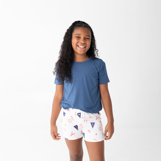 A young girl with long curly black hair stands against a plain white studio background, wearing a short-sleeve blue t-shirt and white shorts with a small multicolor beach pattern.