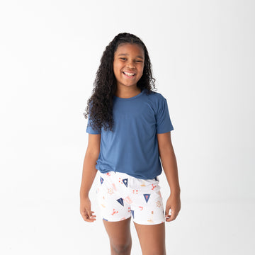 A young girl with long curly black hair stands against a plain white studio background, wearing a short-sleeve blue t-shirt and white shorts with a small multicolor beach pattern.