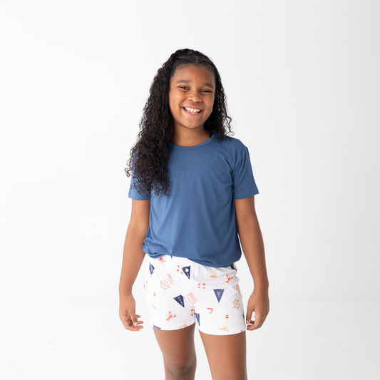 A young girl with long curly black hair stands against a plain white studio background, wearing a short-sleeve blue t-shirt and white shorts with a small multicolor beach pattern.