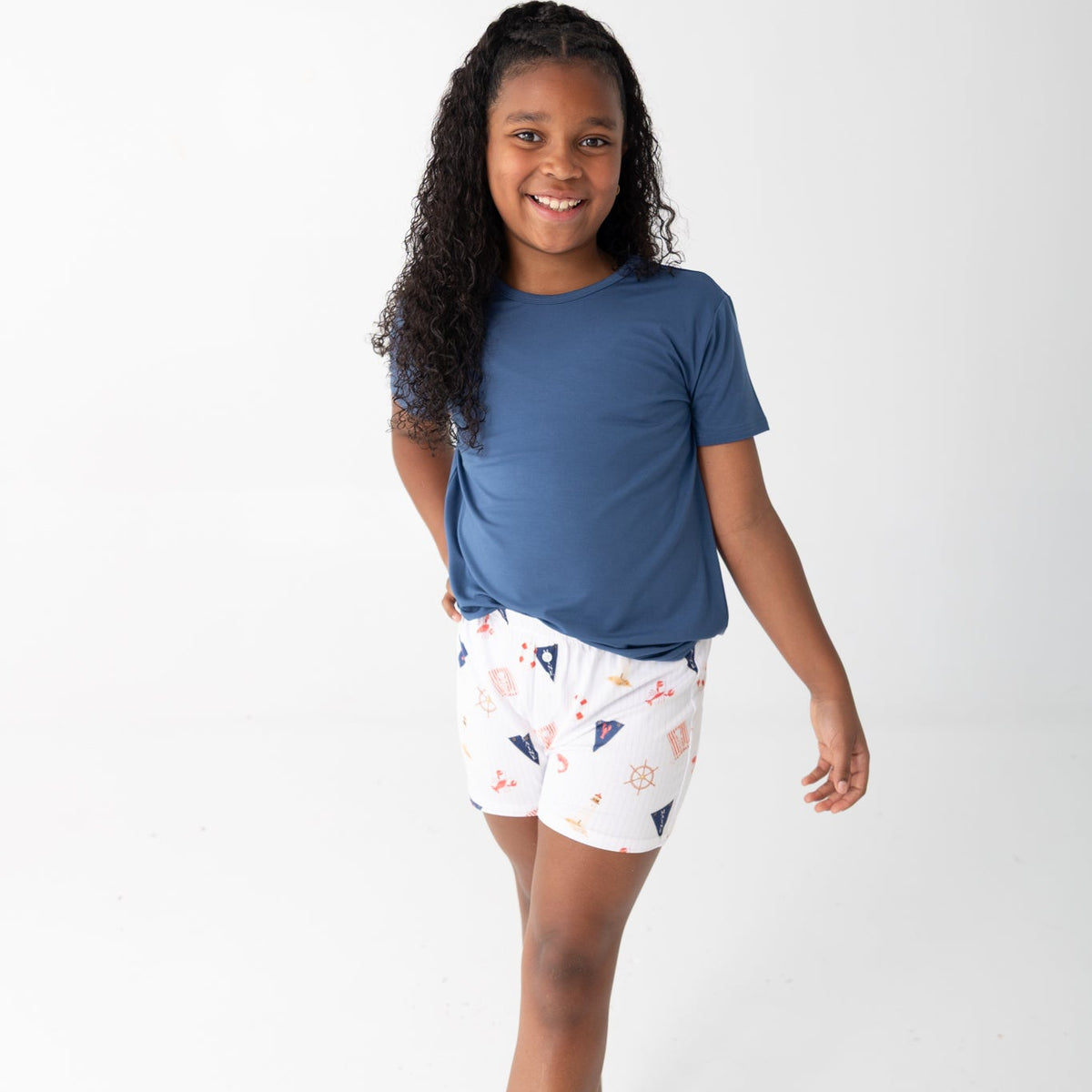 A young girl with long curly dark hair stands barefoot against a plain white studio background, wearing a short-sleeve blue t-shirt and white shorts with a beach nautical print.