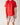 The image shows a child standing against a light-colored wall. They are wearing a plain red short-sleeve t-shirt and white shorts with a colorful pattern. The design on the shorts includes various food items like burgers, fries, and drinks, along with cartoon-style animal faces. The child's arms are relaxed by their sides, and their face is partially out of frame.