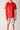 The image shows a child standing against a light-colored wall. They are wearing a plain red short-sleeve t-shirt and white shorts with a colorful pattern. The design on the shorts includes various food items like burgers, fries, and drinks, along with cartoon-style animal faces. The child's arms are relaxed by their sides, and their face is partially out of frame.
