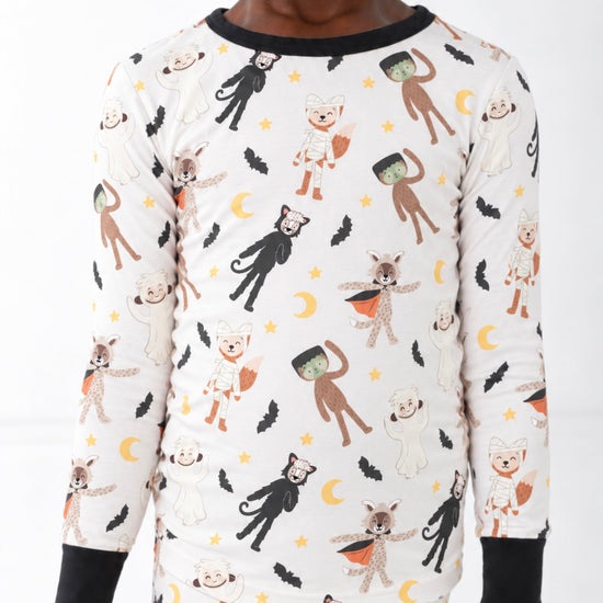 A child wearing a long-sleeve and matching pajama set with an all-over Halloween-themed print of cartoon monsters, cats, dogs, bats, moons, and stars on an off-white background, with black trim at the collar and cuffs.
