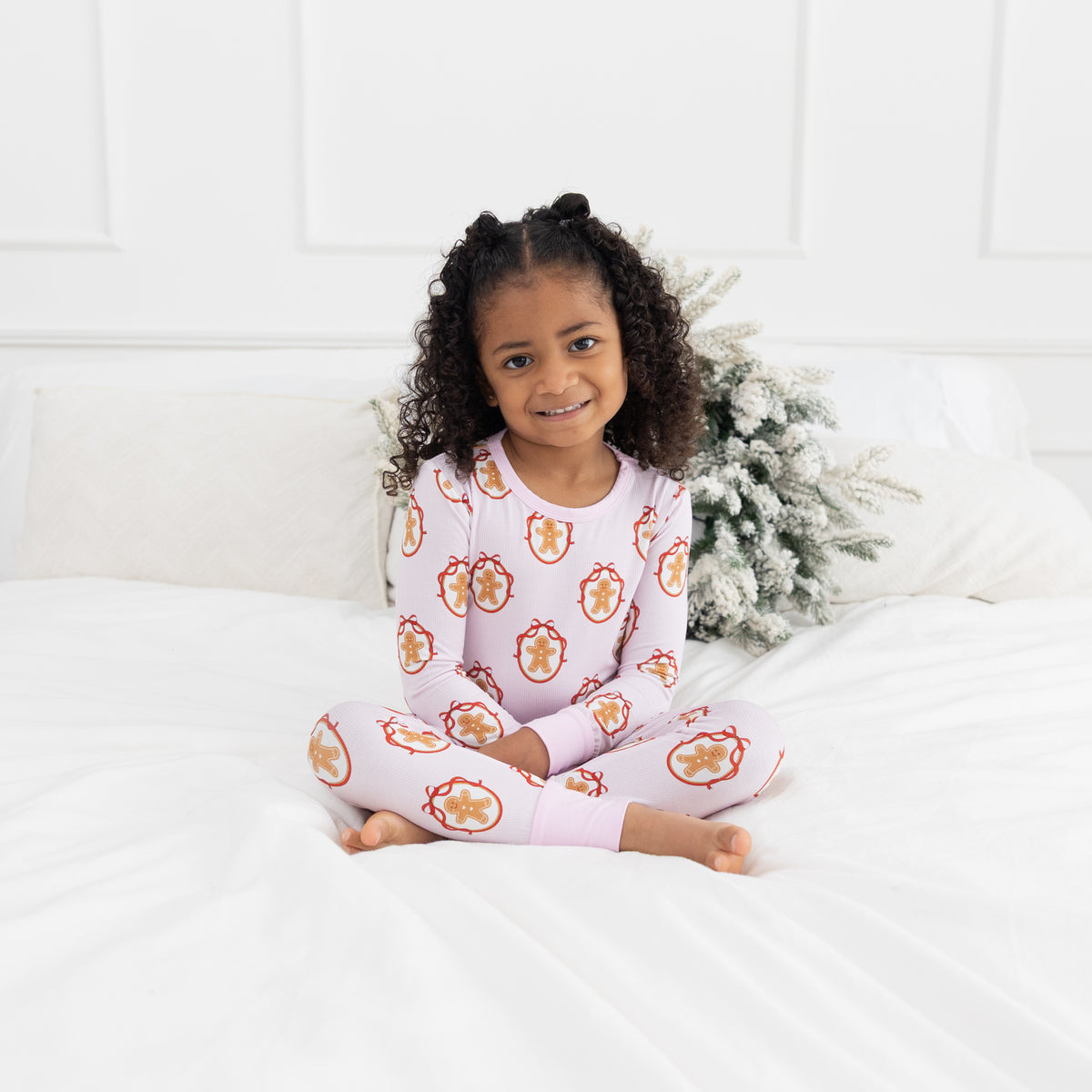 A young girl with curly hair sits cross-legged on a white bed wearing a light pink pajama set with a repeating gingerbread man pattern. A white headboard and a small frosted Christmas wreath are visible in the background.