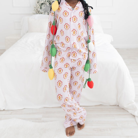 A young girl stands barefoot in a bright bedroom wearing a light pink pajama with a light pink and white stripes long-sleeve pajama set with an allover repeating gingerbread man print. 
