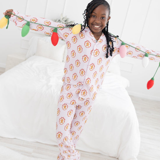 A young girl smiling and posing on a white bed while wearing a light pink pajama with a light pink and white stripes long-sleeve pajama set with an allover repeating gingerbread man print. She is holding a colorful string of Christmas-style lights across her arms in a bright, white bedroom.