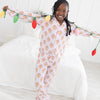 A young girl smiling and posing on a white bed while wearing a light pink pajama with a light pink and white stripes long-sleeve pajama set with an allover repeating gingerbread man print. She is holding a colorful string of Christmas-style lights across her arms in a bright, white bedroom.