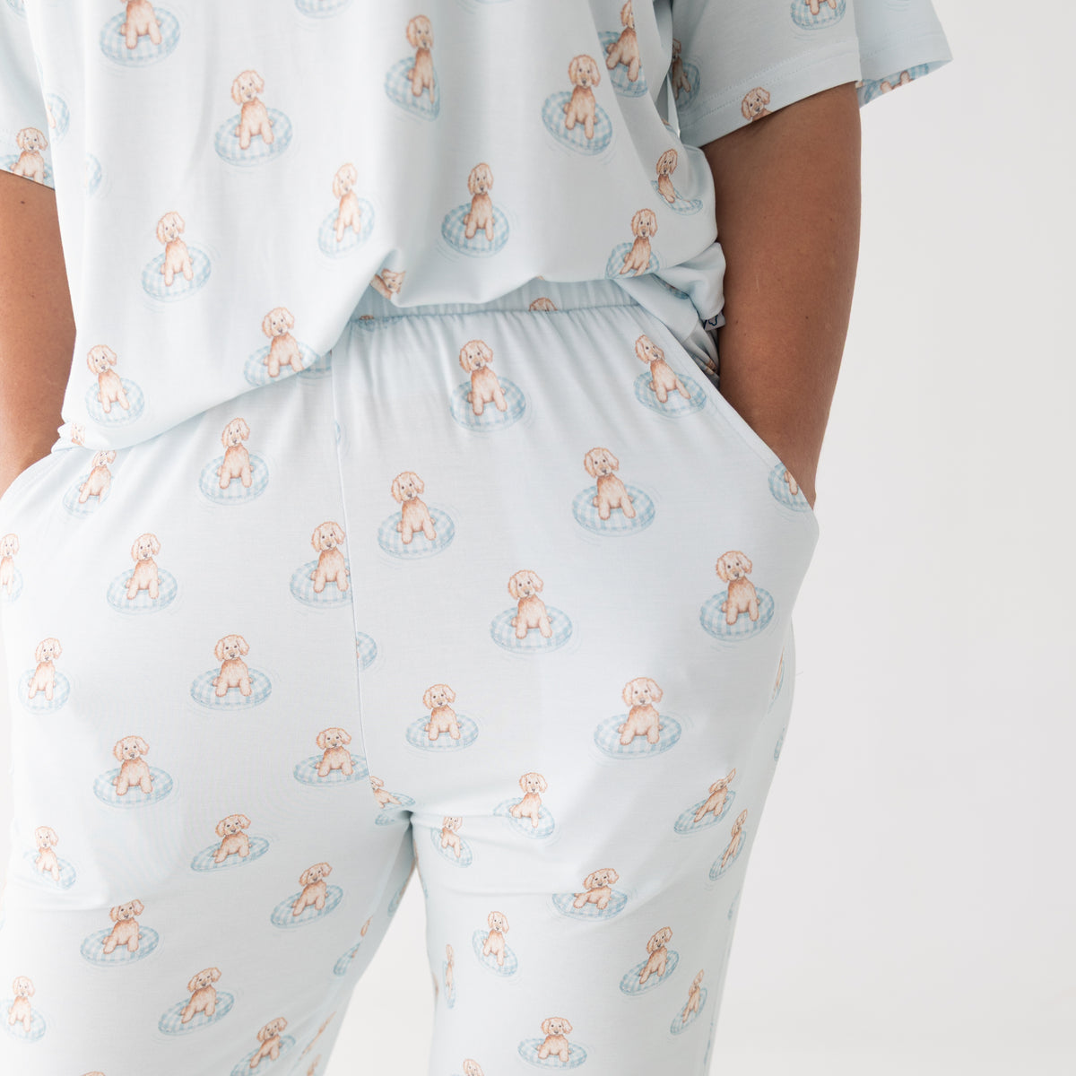 A close-up of a person wearing a white base pajama set with an all-over repeating print of small brown puppy sitting in blue floaty. The top has short sleeves, and the pants have side pockets.