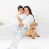 A woman and a young boy wearing matching light blue and white patterned pajamas sit together on a white bed, with a small fluffy tan dog sitting beside them. Pajamas featuring repeating brown puppy in a blue floaty print.