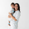 A smiling woman holding a toddler in a matching base pajama set with an all-over repeating print of small brown puppy sitting in blue floaty. The top for the woman has short sleeves, and the pants have side pockets.