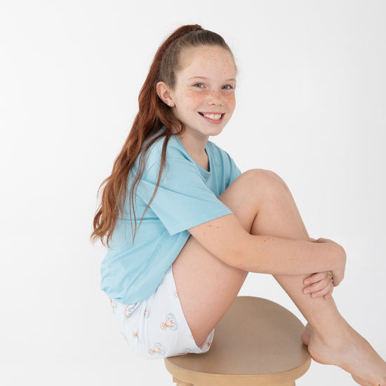 A young girl with freckles sitting on a wooden stool, wearing a light blue t-shirt and white shorts with repeating a brown puppy in a blue floaty print