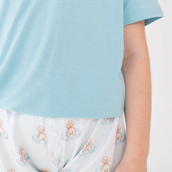 A close-up of a person wearing light blue t shirt with white shorts featuring a repeating a brown puppy in a blue floaty print
