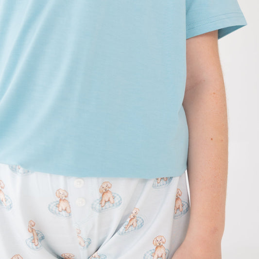A close-up of a person wearing light blue t shirt with white shorts featuring a repeating a brown puppy in a blue floaty print