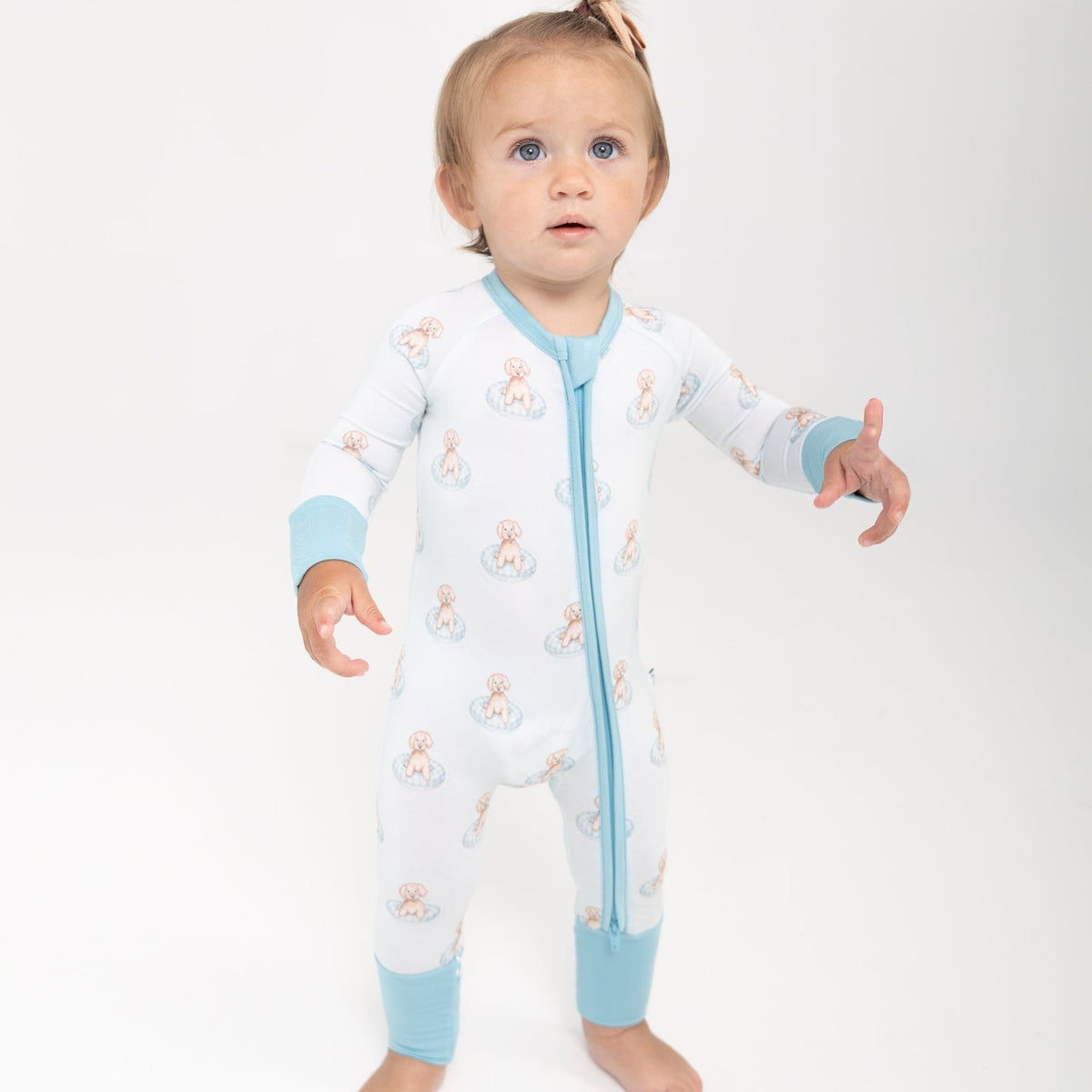 A baby girl wearing a white long-sleeve pajamas with a repeating a brown puppy in a blue floaty print with blue zipper lining and blue cuffs
