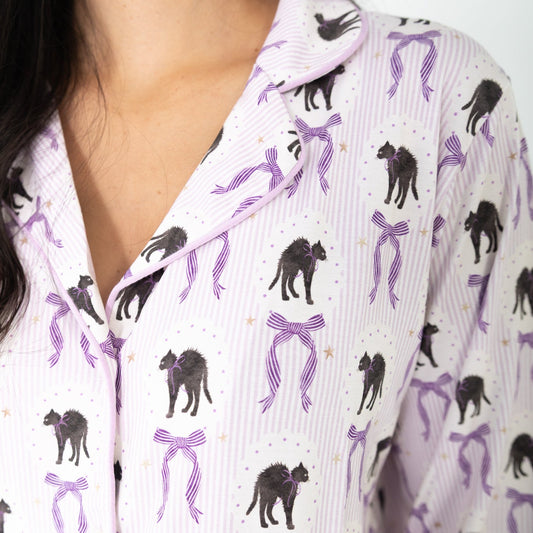 A close-up of a person wearing a pajama top with a white base, light purple vertical stripes, purple ribbon motifs, and black cat illustrations.