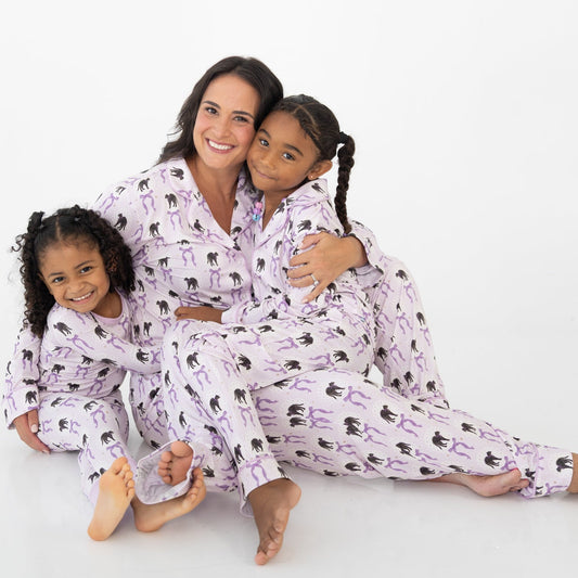 A woman and two young girls sitting together on a white studio floor, wearing matching pajama sets with a white base, purple vertical stripes, and an all-over print of black cat silhouettes and purple bow-tie motifs.