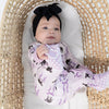 A baby wearing a black bow headband and a light purple footed sleeper with a black cat pattern, lying inside a woven rattan Moses basket.