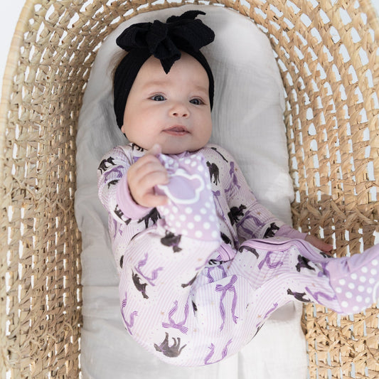 A baby wearing a black bow headband and a light purple footed sleeper with a black cat pattern, lying inside a woven rattan Moses basket.