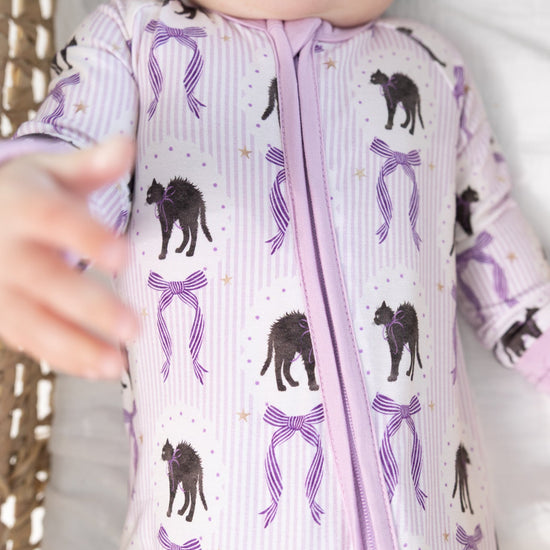 A close-up of a baby wearing a light purple and white striped romper with a purple zipper and an all-over print of black cats  and purple bows.