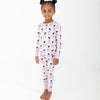 A young girl with her hair in two buns stands barefoot against a plain white studio background, wearing a light purple and white striped two piece long sleeve pajama set featuring an all-over print of black cats and purple bows.