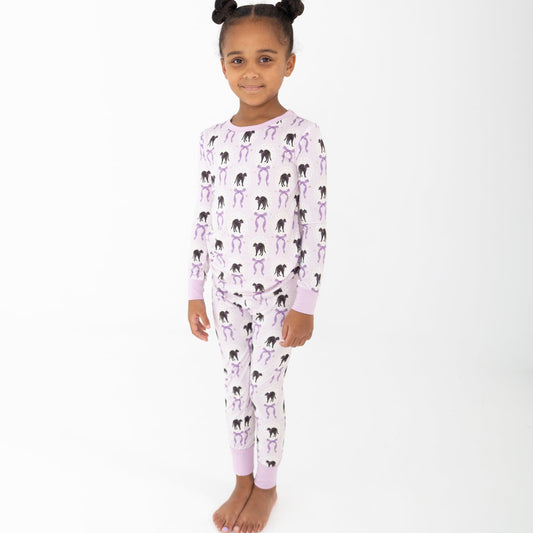 A young girl with her hair in two buns stands barefoot against a plain white studio background, wearing a light purple and white striped two piece long sleeve pajama set featuring an all-over print of black cats and purple bows.