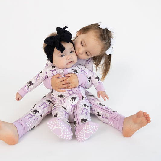 Two young girls sitting on a white studio floor wearing matching light purple pajamas with a black cat print with dainty purple bows. The girl on the right is kissing the girl on the left, who is wearing a large black bow headband.