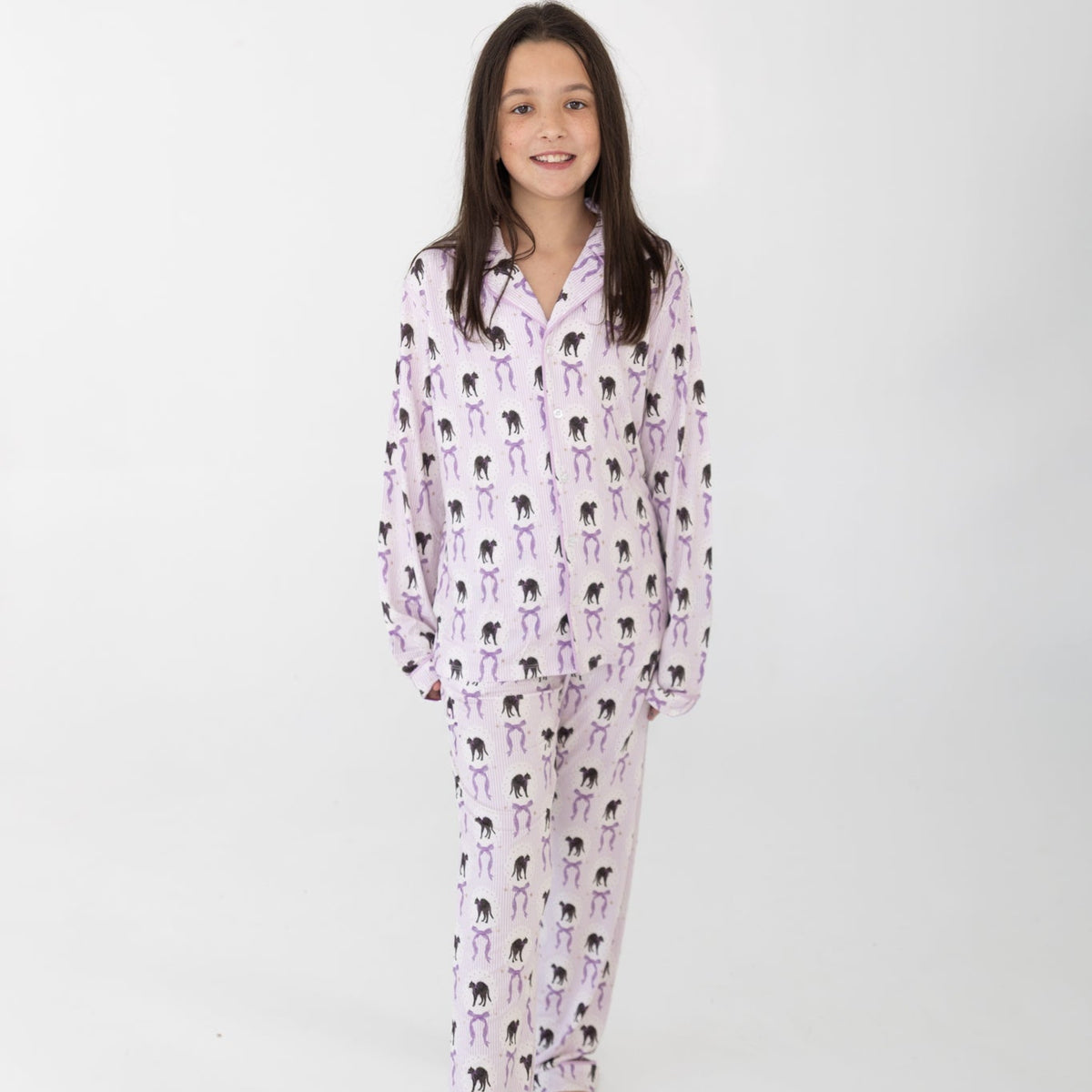 A young girl modeling a two-piece pajama set featuring a light pink and white background with a black cat print with purple bows, standing against a plain white studio background.