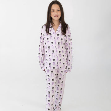 A young girl modeling a two-piece pajama set featuring a light pink and white background with a black cat print with purple bows, standing against a plain white studio background.