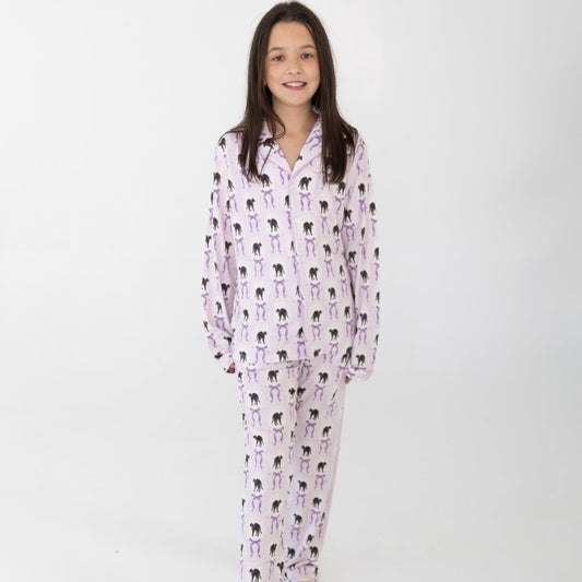 A young girl modeling a two-piece pajama set featuring a light pink and white background with a black cat print with purple bows, standing against a plain white studio background.