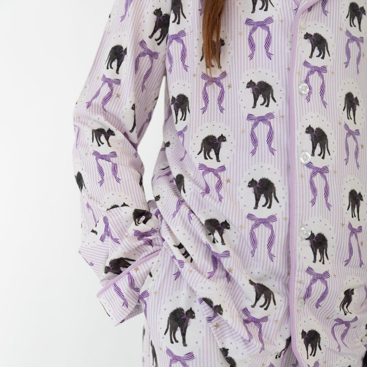 A close-up of a person wearing a matching pajama set with a white base, purple vertical stripes, and an all-over print of black cat silhouettes and purple bow-tie motifs.