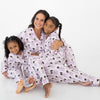 A woman and two young girls sitting together on a white studio floor, wearing matching light purple and white striped two piece long sleeve pajama set featuring an all-over print of black cats and purple bows.