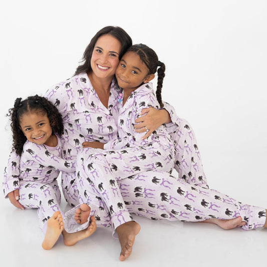 A woman and two young girls sitting together on a white studio floor, wearing matching light purple and white striped two piece long sleeve pajama set featuring an all-over print of black cats and purple bows.