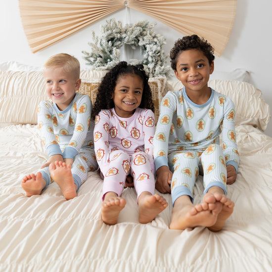 Three children sitting on a bed wearing matching long-sleeve pajamas with a repeating gingerbread pattern. 