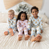 Three children sitting on a bed wearing matching long-sleeve pajamas with a repeating gingerbread pattern. 