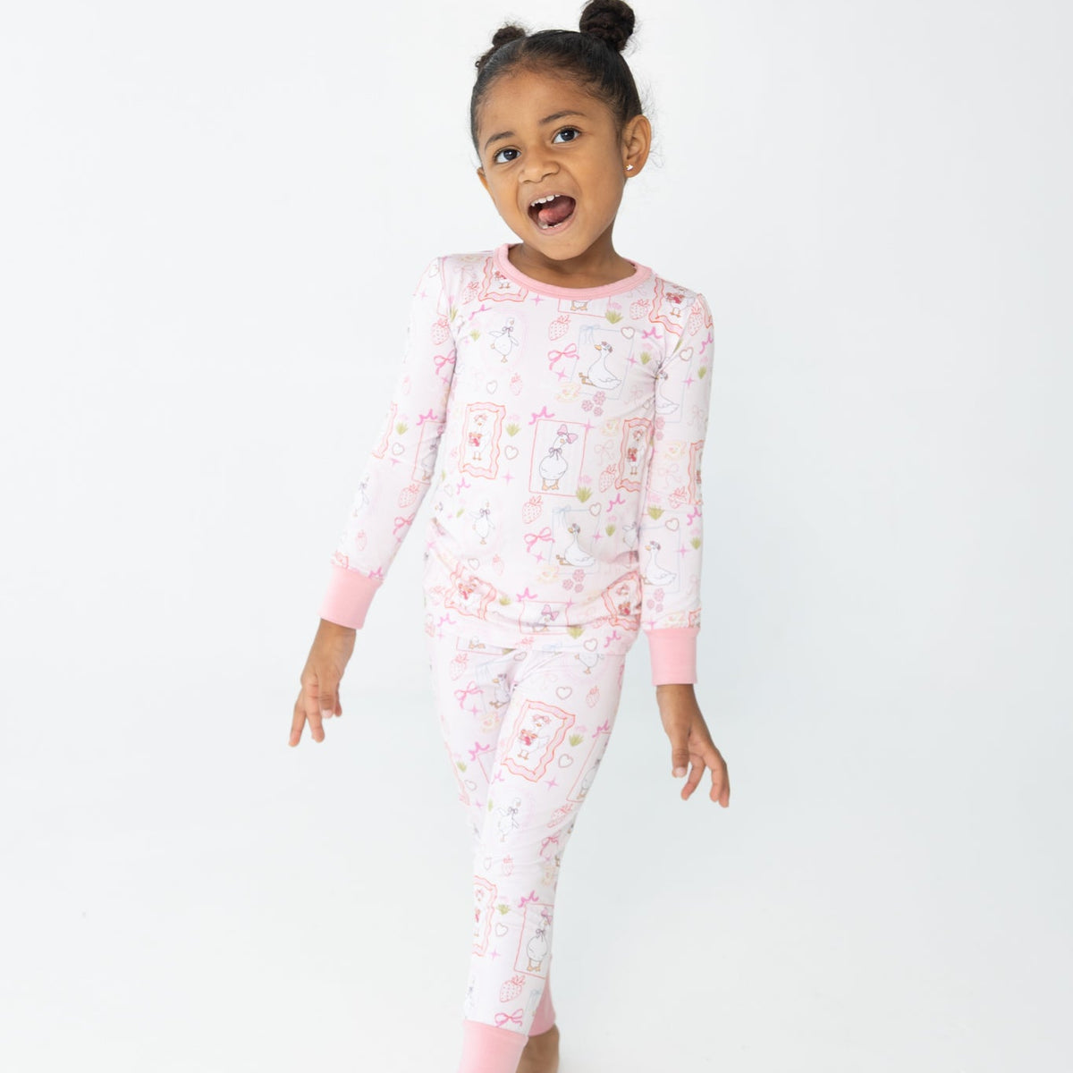 A young girl with her hair in a high bun is wearing a pink and white patterned two piece pajamas featuring silly gooses in a beach theme with strawberries, hearts and pink bows.