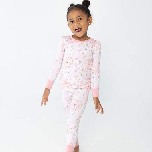 A young girl with her hair in a high bun is wearing a pink and white patterned two piece pajamas featuring silly gooses in a beach theme with strawberries, hearts and pink bows.