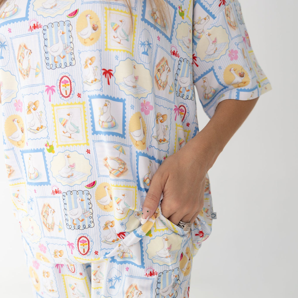 A close-up of a person wearing a matching two-piece pajama set that have blue and white patterned featuring silly gooses in a beach theme with watermelon, beach theme and palm trees.