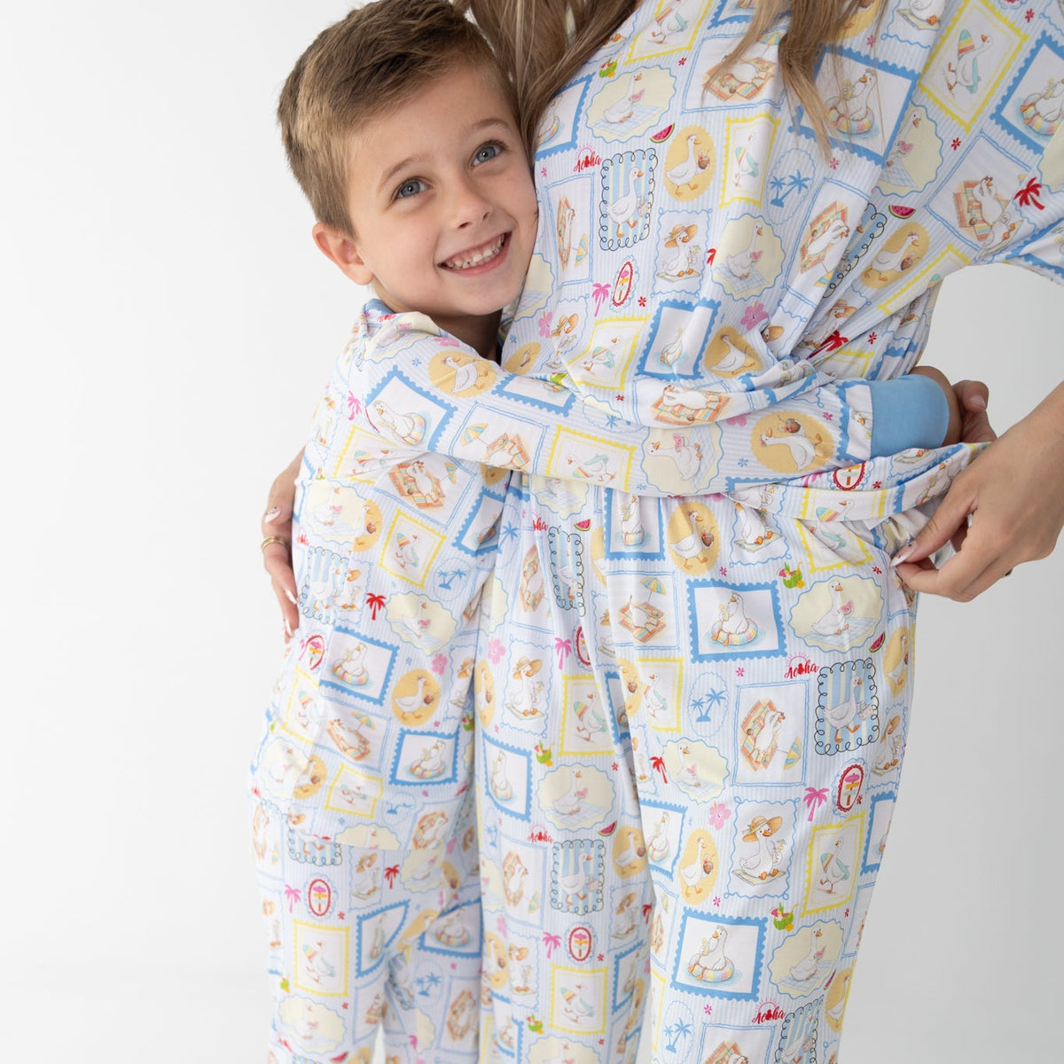 A smiling young boy in a long sleeve two piece pajama set hugs a woman wearing matching pajamas short sleeve shirt with pants. The pajama sets have blue and white patterned featuring silly gooses in a beach theme with watermelon, beach theme and palm trees.