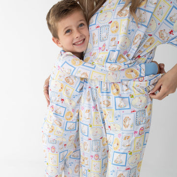 A smiling young boy in a long sleeve two piece pajama set hugs a woman wearing matching pajamas short sleeve shirt with pants. The pajama sets have blue and white patterned featuring silly gooses in a beach theme with watermelon, beach theme and palm trees.