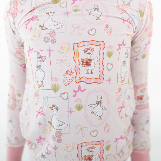 A close-up of a long-sleeve toddler long sleeve and pants two piece set featuring a cute cartoon print of white ducks, strawberries, bows, and hearts on a light pink background.