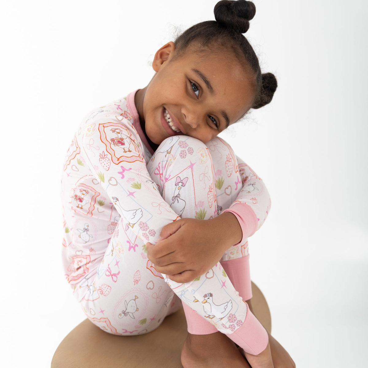 A young girl with her hair in a high bun is wearing a pink and white patterned two piece pajamas featuring silly gooses in a beach theme with strawberries, hearts and pink bows.