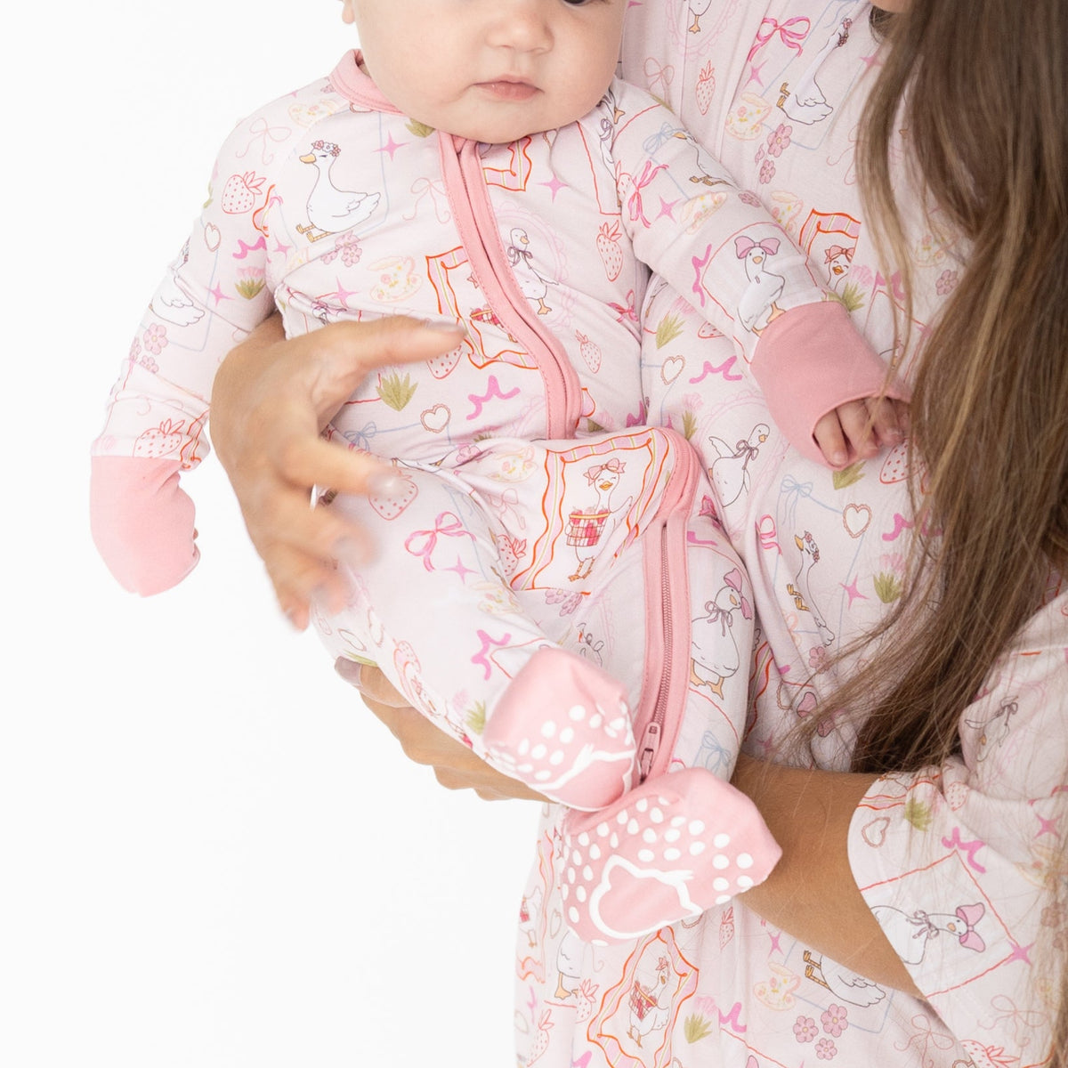 A baby wearing a pink and white patterned zippy with a pink zipper featuring silly gooses, strawberries, hearts and bows. Woman is wearing a matching short sleeve shirt with pants set.