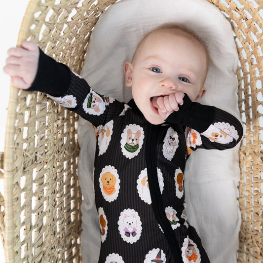 A baby wearing a black long-sleeve zippy romper with dogs dressed up in costumes, sitting inside a woven rattan bassinet.