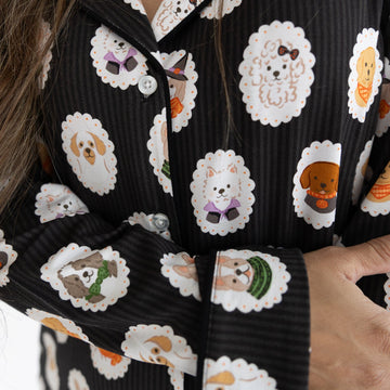 Close-up of a person wearing black pajama long-sleeve top with a white dog print pattern. The pajama top features a button-down front and a cozy collar, with a repeating pattern of dogs dressed up in costumes. 