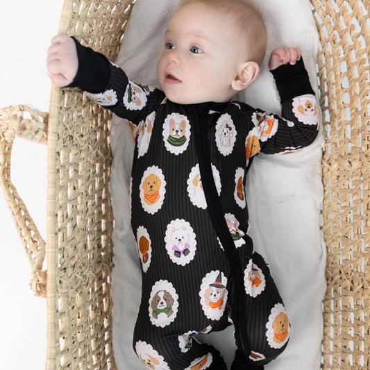 A baby lying on a white cushion inside a woven natural fiber Moses basket, wearing a black long-sleeve zippy romper with dogs dressed up in costumes, sitting inside a woven rattan bassinet.